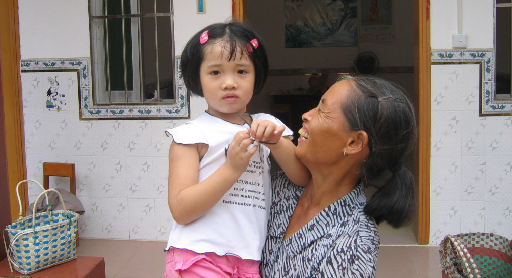 A young girl looks at the camera while being embraced by her grandmother while visiting her village in China.
